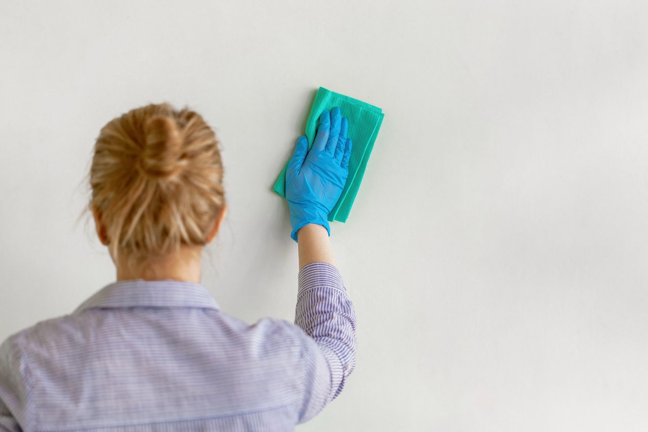 Employee hand in blue rubber protective glove wiping wall from dust with dry rag. General or regular cleanup. Commercial cleaning company. Copy space.