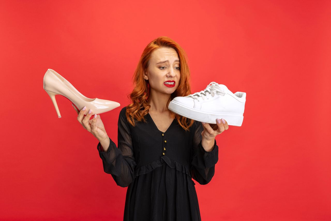 Portrait of expressive woman holding sneakers and heeled shoes isolated on red studio background