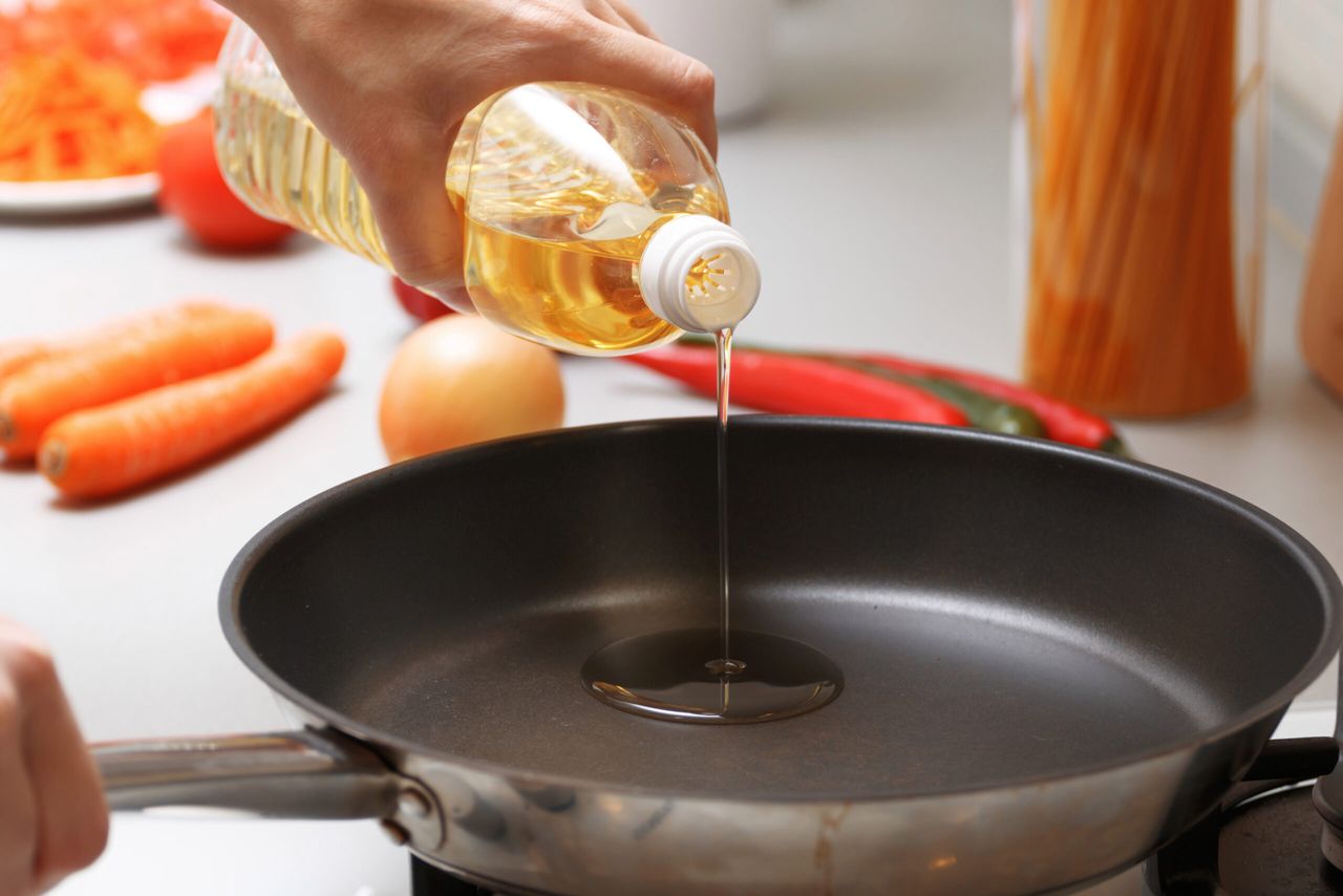 A woman pouring oil from a bottle into the pan in the kitchen, near fresh vegetables and pasta.