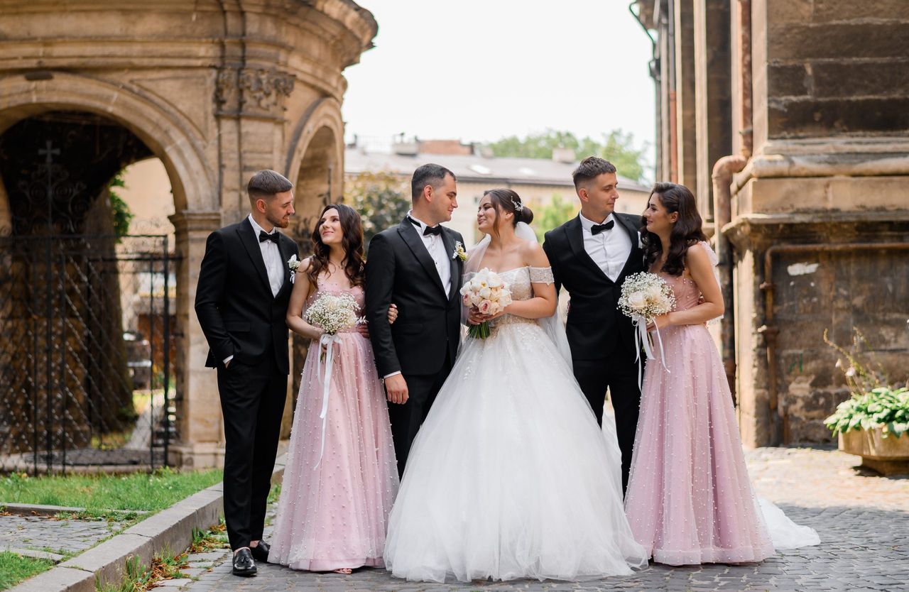 Front view of beautiful bride and groom in wedding apparel, smiling and looking to each other while standing between friends. Pretty bridesmaids with curly long hair, dressed in pink evening gowns, holding flowers arrangement, standing near elegant guys in suit and posing on ancient city background