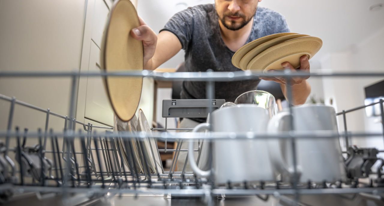 A man in front of an open dishwasher takes out or puts down dishes.