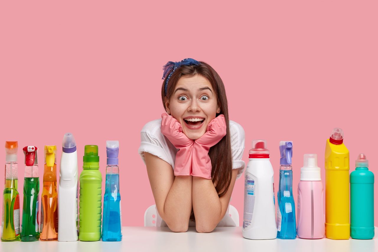 Happy young Caucasian woman holds chin, wears rubber protective gloves and headband, smiles broadly, surrounded with bottles of detergents, isolated over pink background. Female janitor indoor