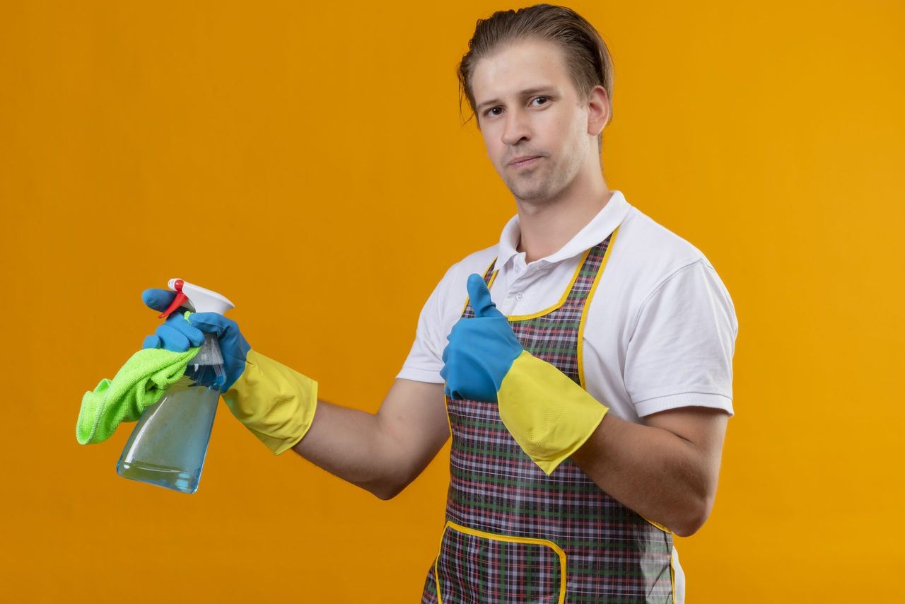 young hansdome man wearing apron and rubber gloves holding cleaning spray and rug looking at camera with confident smile on face showing thumbs up standing over orange background