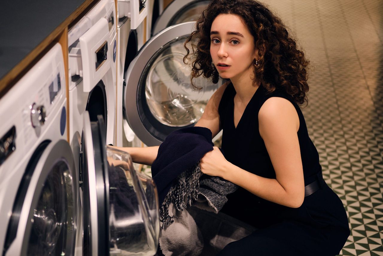 Portrait of brunette girl thoughtfully looking in camera taking out clothes from washing machine in self-service laundry