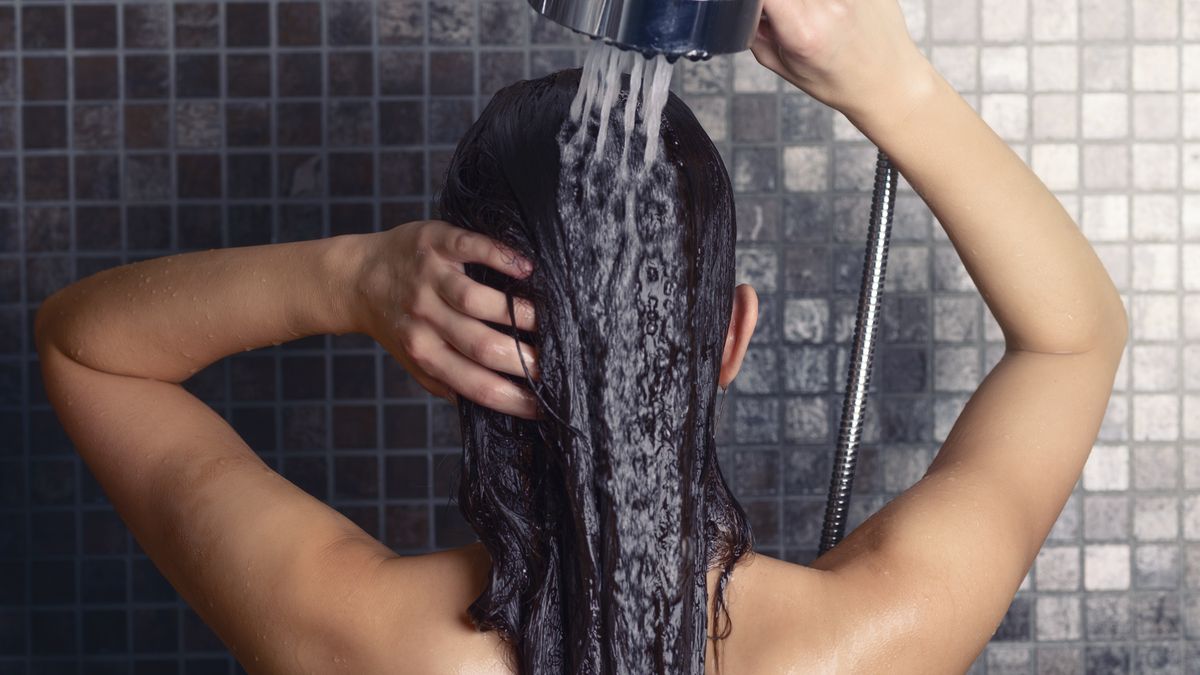 Young woman washing her long hair under the shower standing with her back to the camera rinsing it off under the jet of water with her head partially turned to the side, over grey mosaic tiles