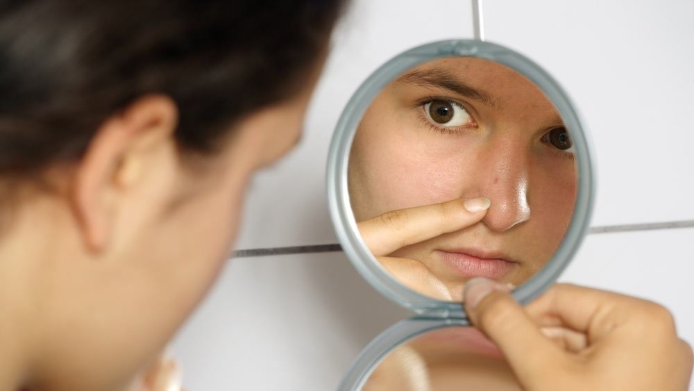 Young teenage female holding a mirror looking at her pimple with concern.