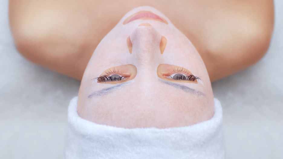 The cosmetologist for procedure of cleansing and moisturizing the skin, applying a sheet mask to the face of a young woman in beauty salon.Cosmetology and professional skin care.