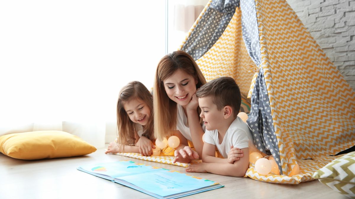 Nanny and little children reading book in tent at home