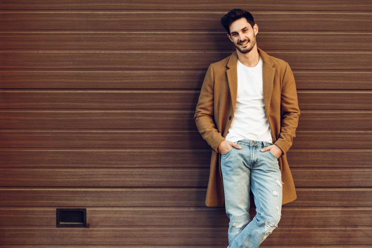Young smiling man wearing demi-season clothes in the street. Young bearded guy with modern hairstyle with coat, blue jeans and t-shirt against urban wooden blinds. 