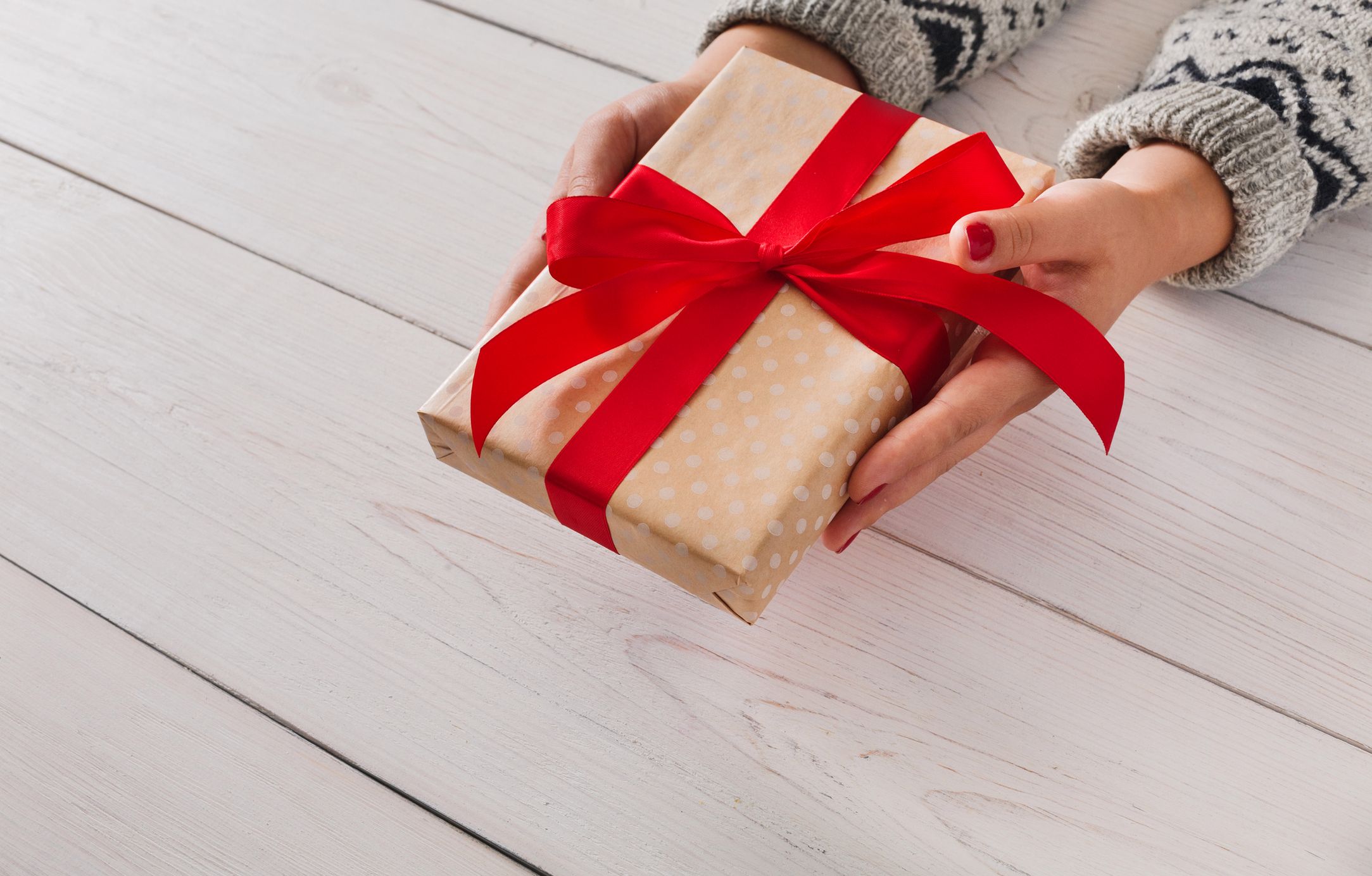 Woman's hands give wrapped christmas or other holiday handmade present in paper with red ribbon. Present box, decoration of gift on white wooden table 