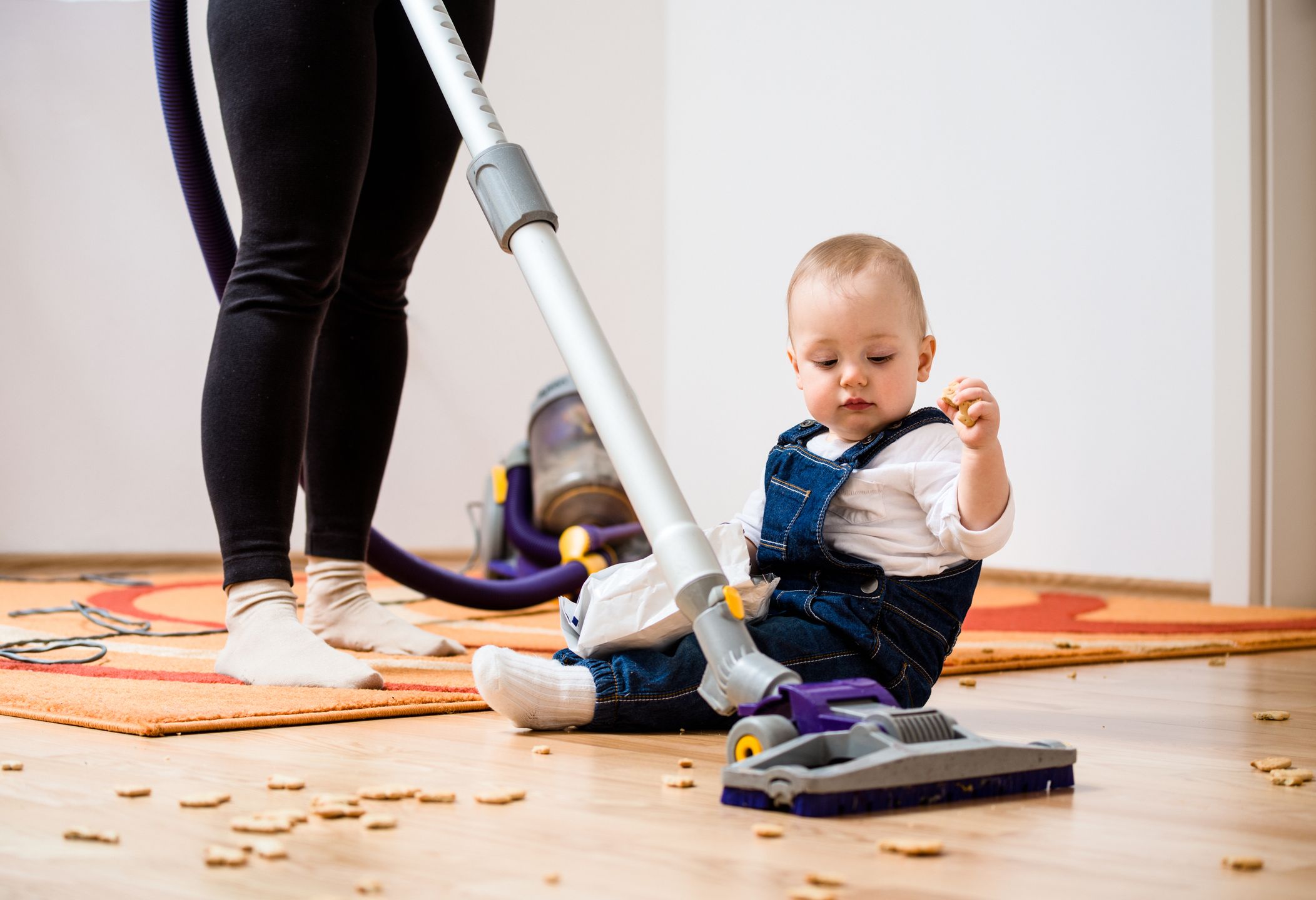 Woman cleaning with vacuum cleaner, baby sitting on floor and biscuits all around 